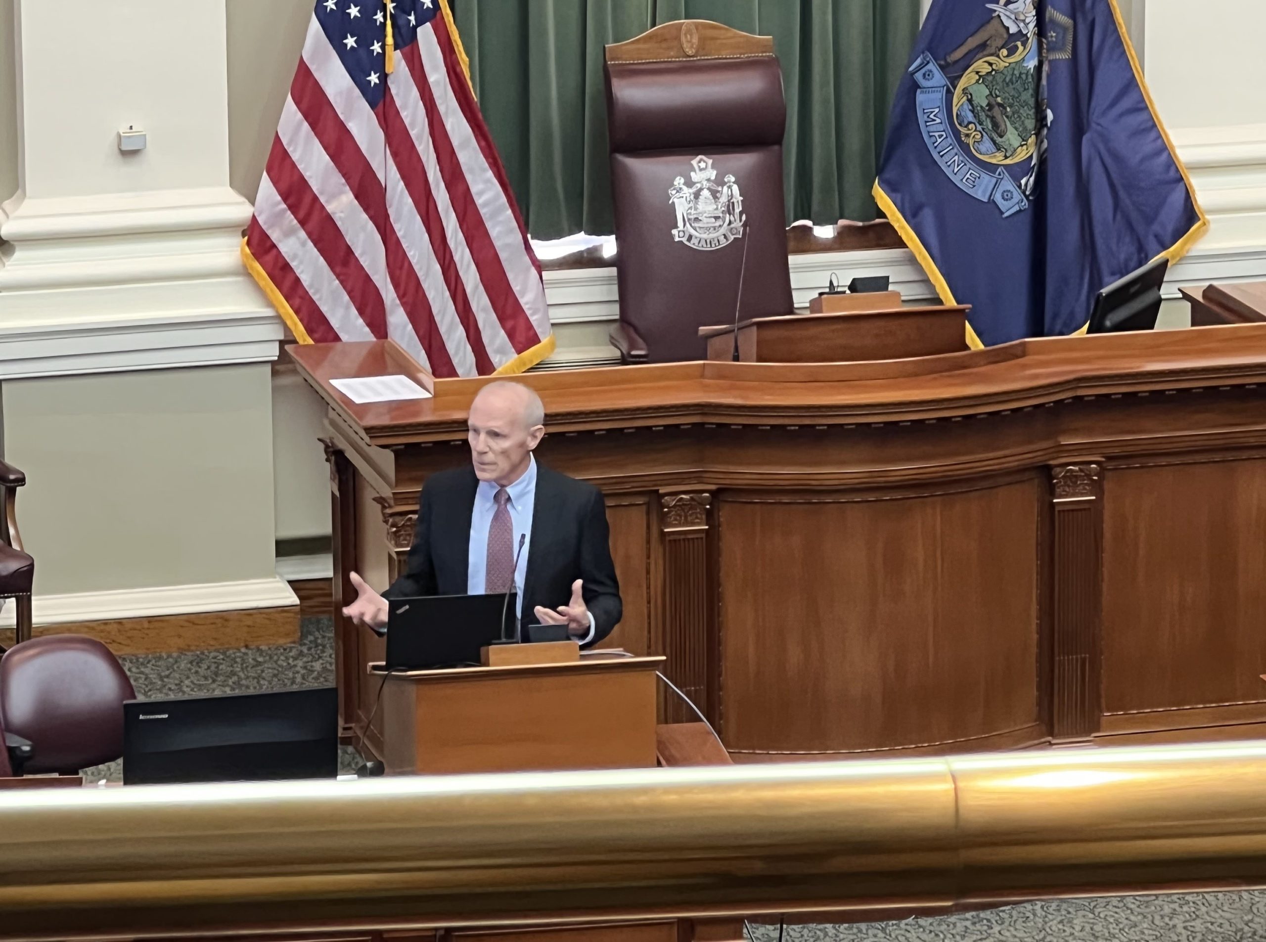 image of man at podium speaking with american and maine flags behind him