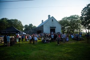 Large crowd gathers for an event outside at sunset in front of a large white barn.