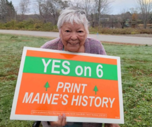 Passamaquoddy Elder Theresa Downing holding orange and green lawn sign with text Yes on 6 Print Maine's History