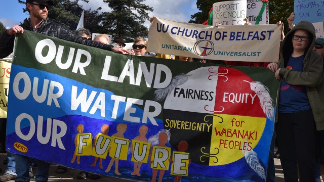 Photo: Anne Henshaw People holding banner painted green and blue with text our land our water our future and circle painted white, red, black and yellow with text fairness equity sovereignty for wabanaki peoples